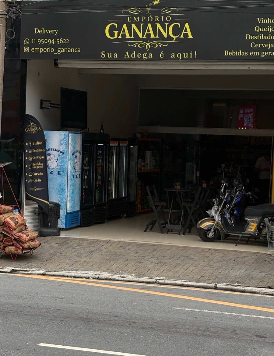 Fachada do Empório Ganança, com o letreiro preto e dourado no alto, geladeiras de bebidas e mesas na entrada aberta para a calçada.