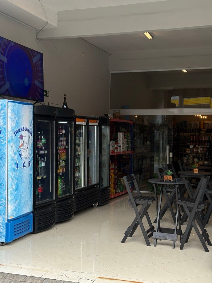 Entrada do Empório Ganança vista da calçada: geladeiras de bebidas à esquerda, mesas altas de madeira escura à direita e, ao fundo, a divisória de vidro que mostra o interior iluminado da loja.