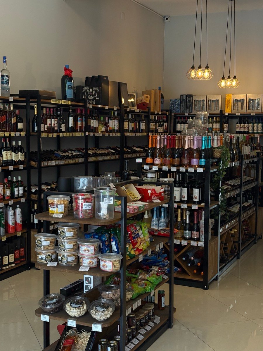 Área de produtos do Empório Ganança: estantes de metal preto e madeira com garrafas de vinho e outras bebidas, potes de petiscos em primeiro plano e luminárias pendentes acesas.